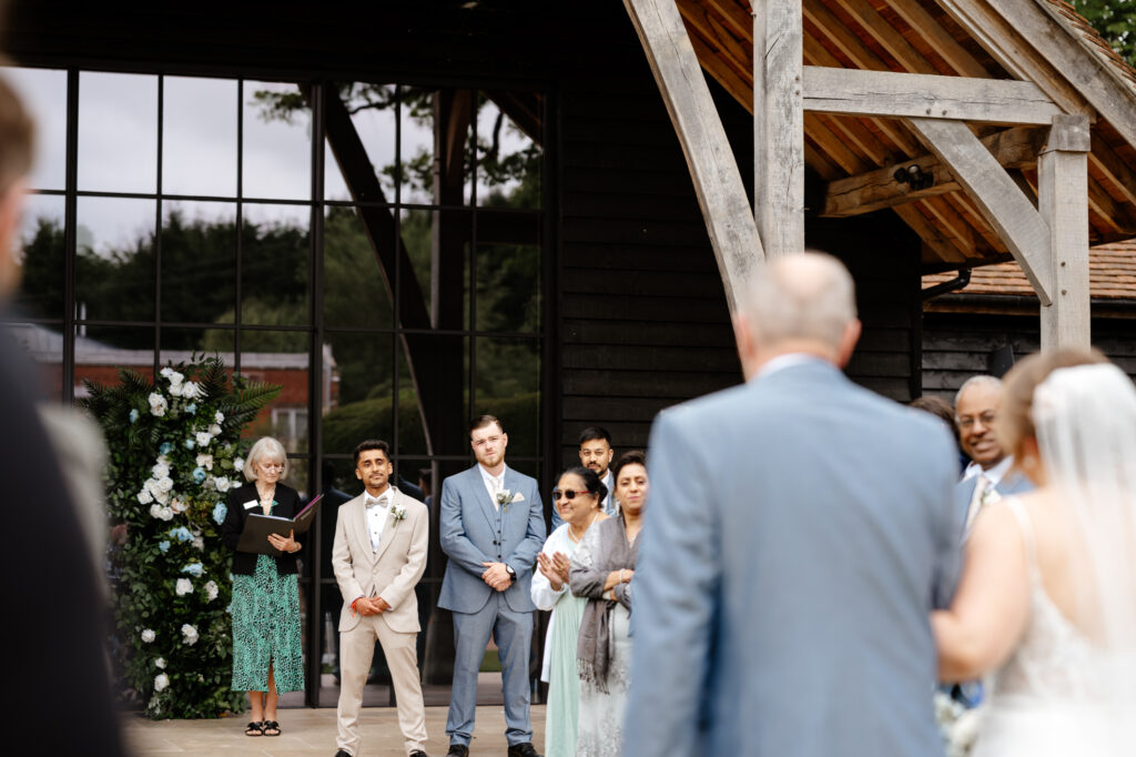 The groom watches with emotion as the bride walks down the aisle with her escort, surrounded by loved ones and a floral backdrop at The Post Berkshire