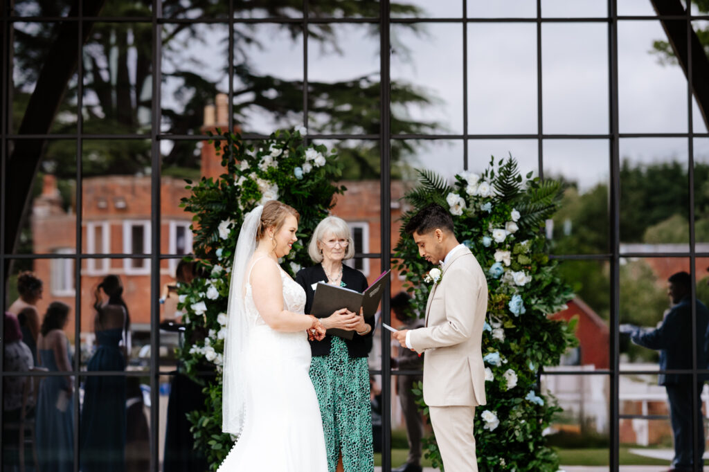 The bride and groom stand facing each other during their outdoor wedding ceremony, exchanging vows in front of a floral arch of white and green blooms. The officiant stands between them holding a folder, while reflections of guests and the venue’s brick buildings appear in the large glass windows behind them.