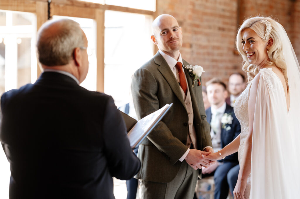Bride in an elegant lace gown with cape sleeves and groom in a green suit with rust-colored tie exchange vows during an intimate indoor ceremony with exposed brick walls and natural light.